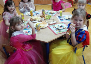 Dzieci w kolorowych strojach siedzą i jedzą poczęstunek.